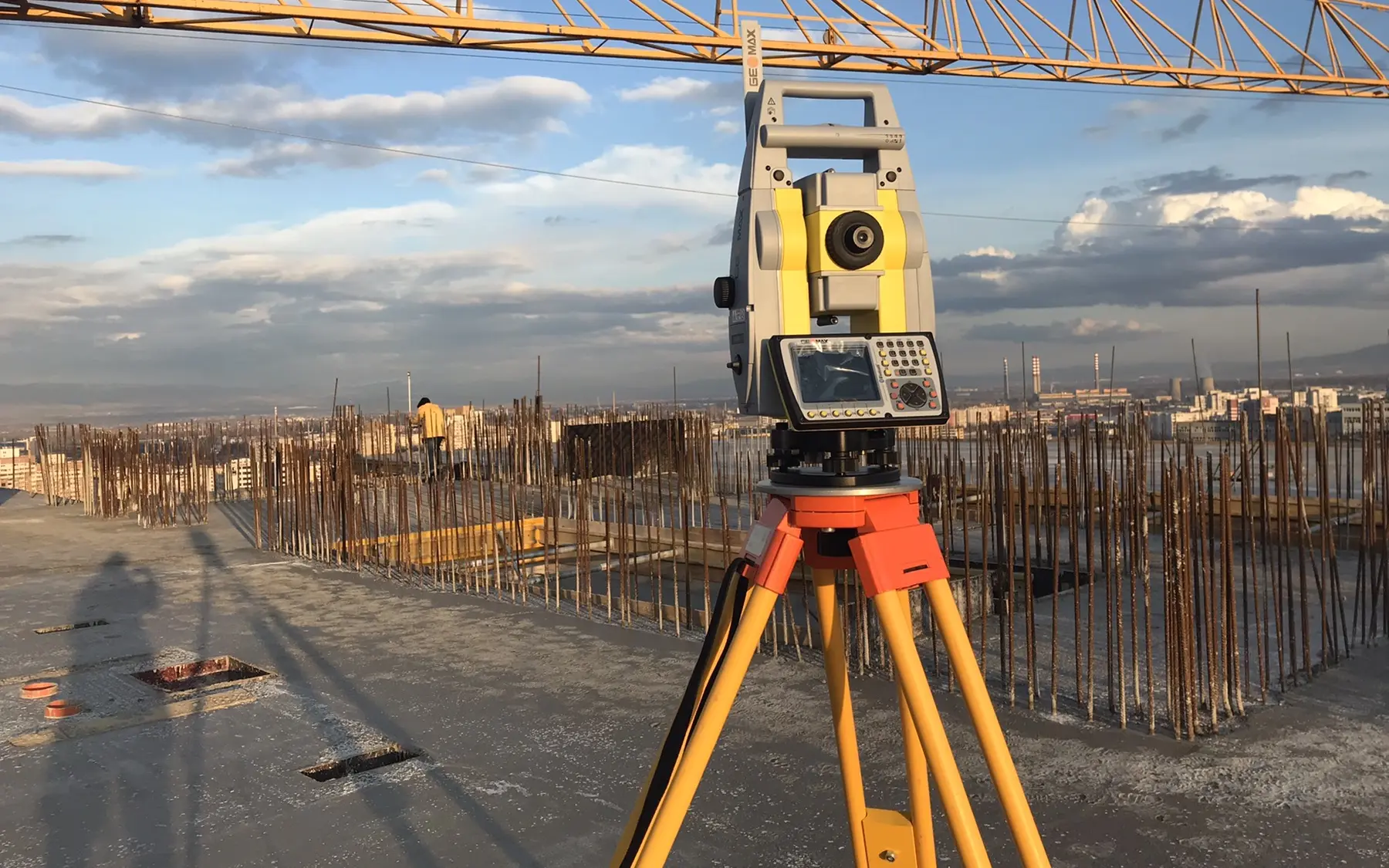 Роботизирана тотална станция GeoMax Zoom на статив върху плоча на строеж, с арматура и кулокран на фона на София